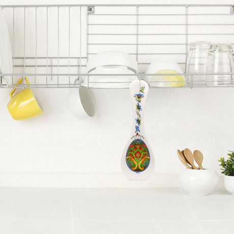 The Green Norwegian Rosemaling Ceramic Spoon Rest, featuring a colorful floral design, sits on a white kitchen counter under a drying rack with cups and bowls, adding Scandinavian charm to your decor.