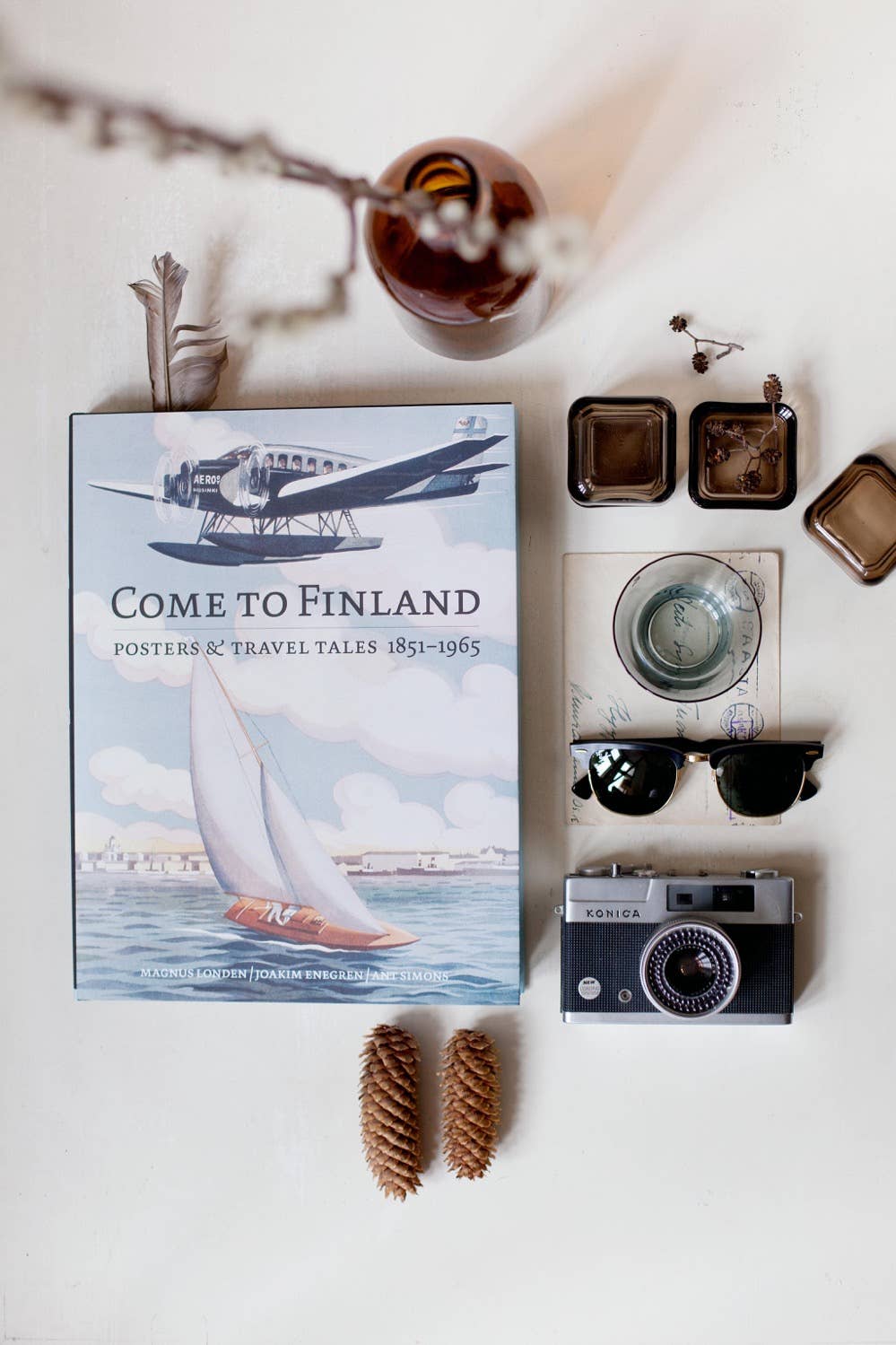 A flat lay featuring the Book: Come to Finland, Coffee Table Book in English (Hardcover), a vintage camera, sunglasses, glass bottles, a postcard, and pine cones on a light surface.