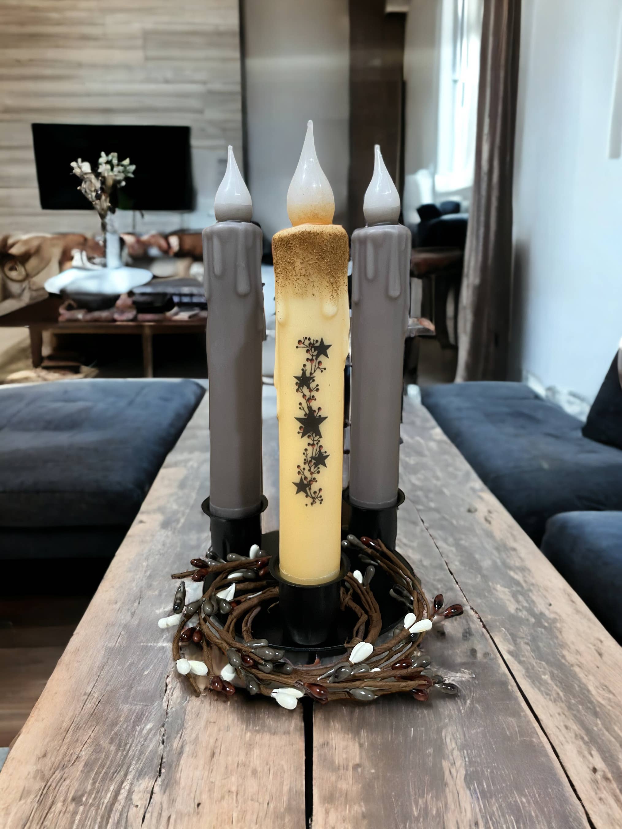 Three decorative candles, two gray and one ornate yellow, are elegantly arranged on the living room centerpiece using the Round Triple Wrought Iron Taper Holder. A wreath encircles their bases atop a wooden table, with the sofa and TV creating a cozy backdrop. The timeless design exudes the charm characteristic of Lancaster blacksmith products.