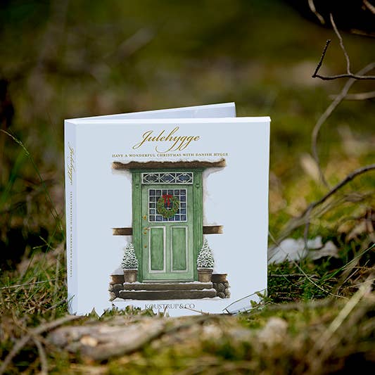 The Card Pack: Julehygge by Marianne Scheel showcases a cover illustration of a green door decorated with a Christmas wreath, set against an outdoor grassy backdrop that captures the warmth and charm of holiday traditions.