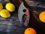 An Alaskan Ulu Style Blade kitchen knife with a red and black diamond wood handle rests on a wooden surface beside three lemons and two oranges. The handmade blade is crafted from Damascus steel.