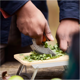 A person chops fresh cilantro on a small wooden cutting board using an Alaskan Ulu Style Blade.