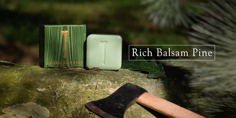 The Balsam Pine Soap, showcased with a T logo embossed on it, is perched on a log beside its box labeled "Rich Balsam Pine body bar." Nearby, an axe complements the scene, set against a backdrop of blurred green foliage that evokes the essence of winter pine forests.