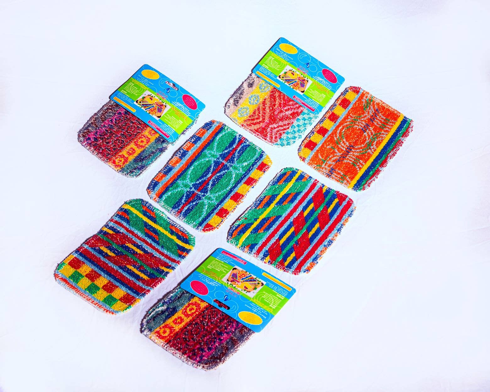 Seven colorful Scrub: Kitchen Scrubber Scrub-Wow pads with packaging are arranged on a white background. Each non-toxic scrubber has a unique multicolored geometric pattern and is safe for Teflon surfaces.
