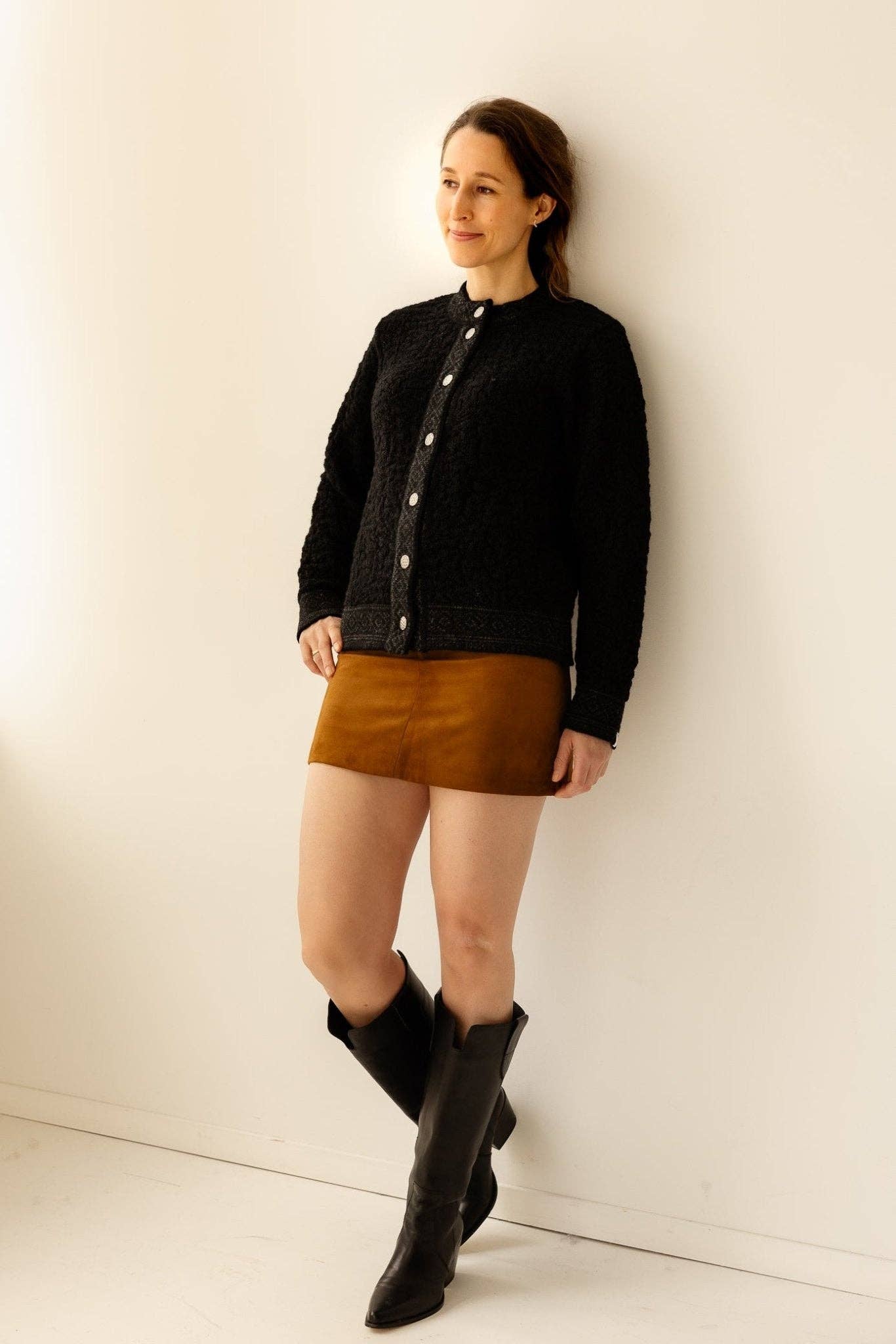 A woman stands against a white wall wearing a white Bryggen Women's Norwegian Cardigan, a brown mini skirt, and black knee-high boots.