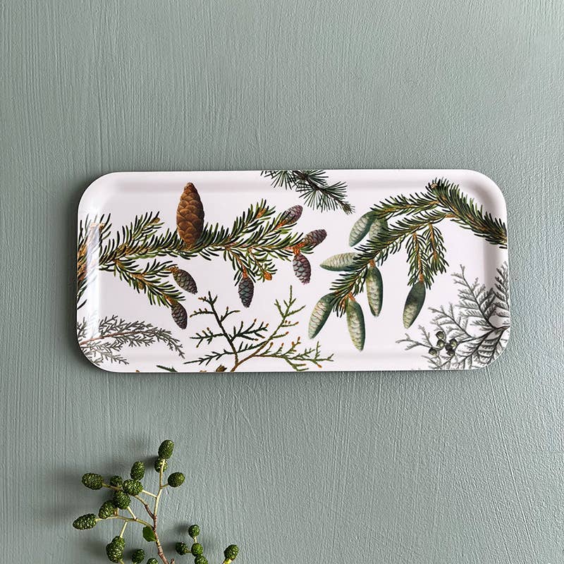 The "Tray: Pine Trees" is a 6x12 inch wood serving tray made in Sweden for Koustrup Co, featuring pine branch and cone botanical art on Nordic birch veneer, shown on a green surface with sprigs of greenery.