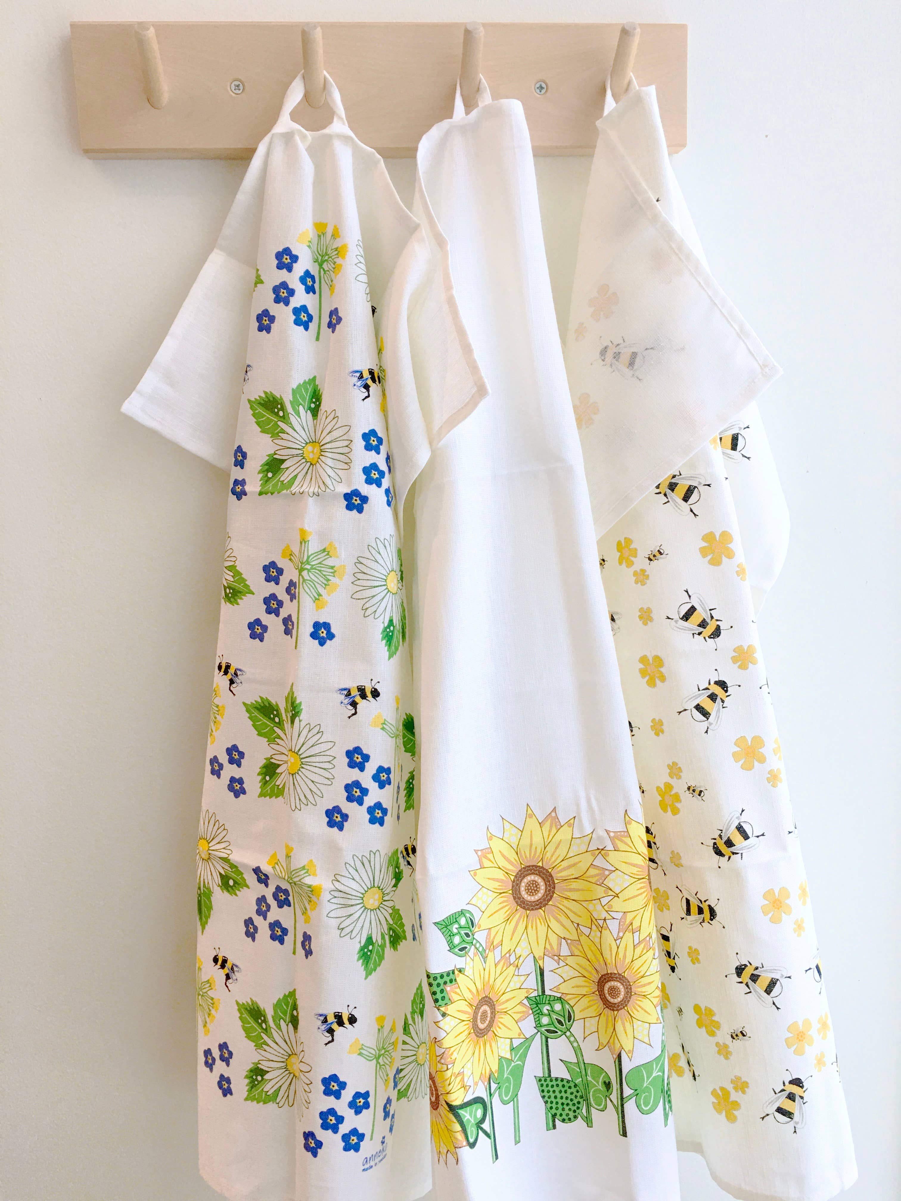 A gift duo featuring one Swedish tea towel and one dish cloth, each hanging on wooden hooks, showcases floral and bee patterns in blue, green, and yellow.