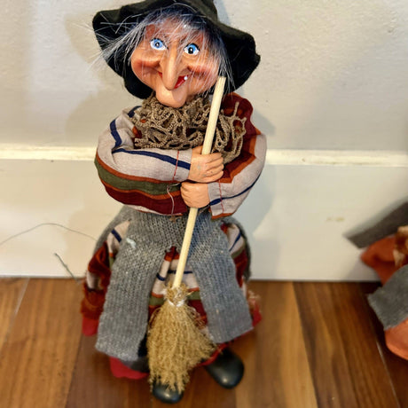 The Marketplace24 Kitchen Witch: Nonna Red, a 10" tall figurine with broom, features a large nose, wide smile, blue eyes, striped clothes, and gray apron—an enchanting Scandinavian good luck charm for your Nordic kitchen décor.