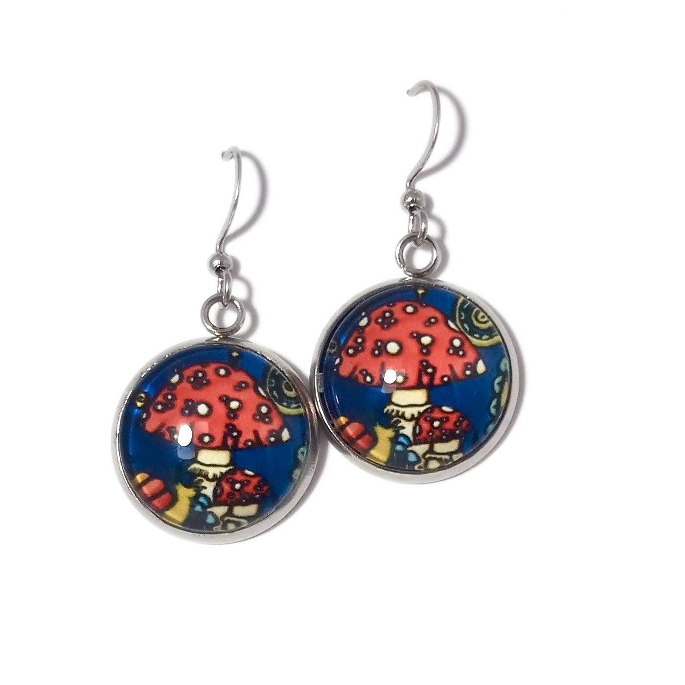 These handmade earrings, known as "Earrings: Mushrooms by Sarah Angst," feature a whimsical mushroom design with red caps and white spots set against a blue background.