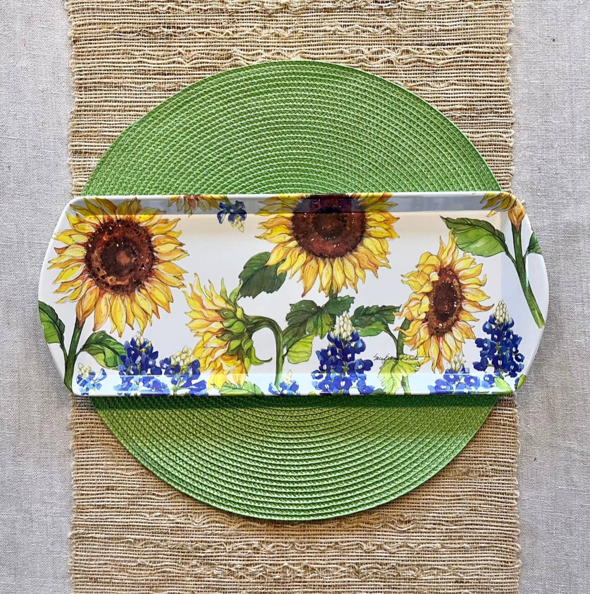 The Tray: Sunflower & Bluebonnets Floral Loaf Serving Charcuterie Tray, with charming sunflower and bluebonnet designs, sits on a green woven placemat atop a beige runner. This eco-friendly bamboo tray adds elegance to any table setting.