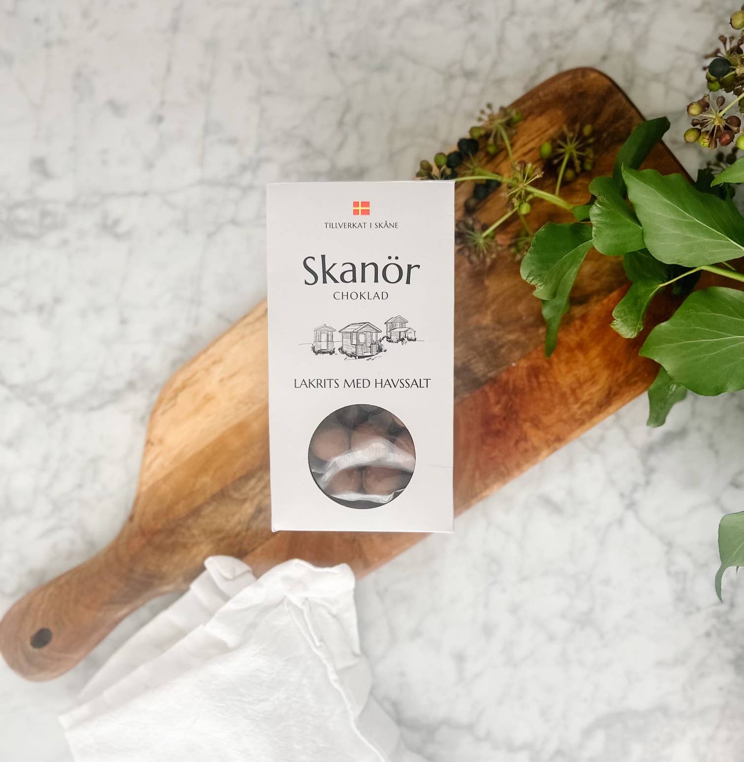 A box of Candy: Skanör - Chocolate Coated Licorice with Sea Salt (100g) rests on a wooden cutting board, surrounded by lush greenery and a white cloth atop a marble surface, capturing the essence of Skåne, Sweden.