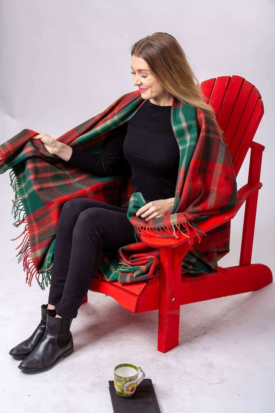 A woman sits on a red wooden chair, wrapped in a New Brunswick Deluxe Highland Tartan Blanket (100% wool, 69" x 62"), with a mug and black book resting on the floor nearby.