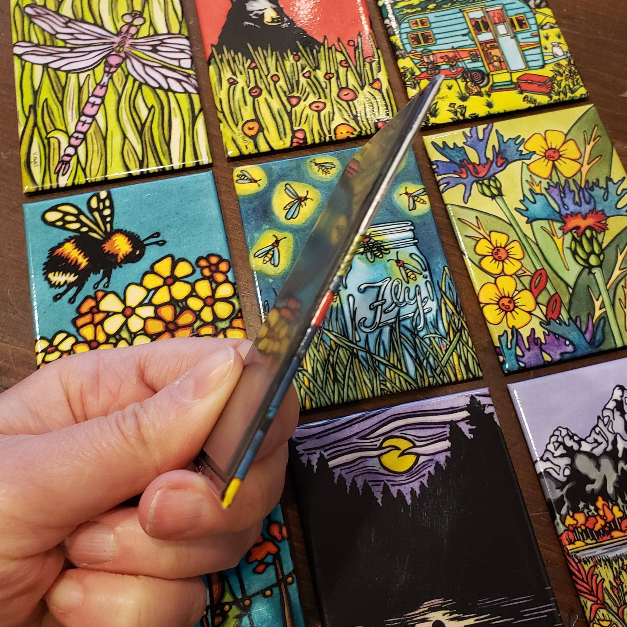 A hand holds a painted tile, surrounded by colorful tiles featuring nature designs and insects, resembling unique-shaped Mushroom Magnets on a table.