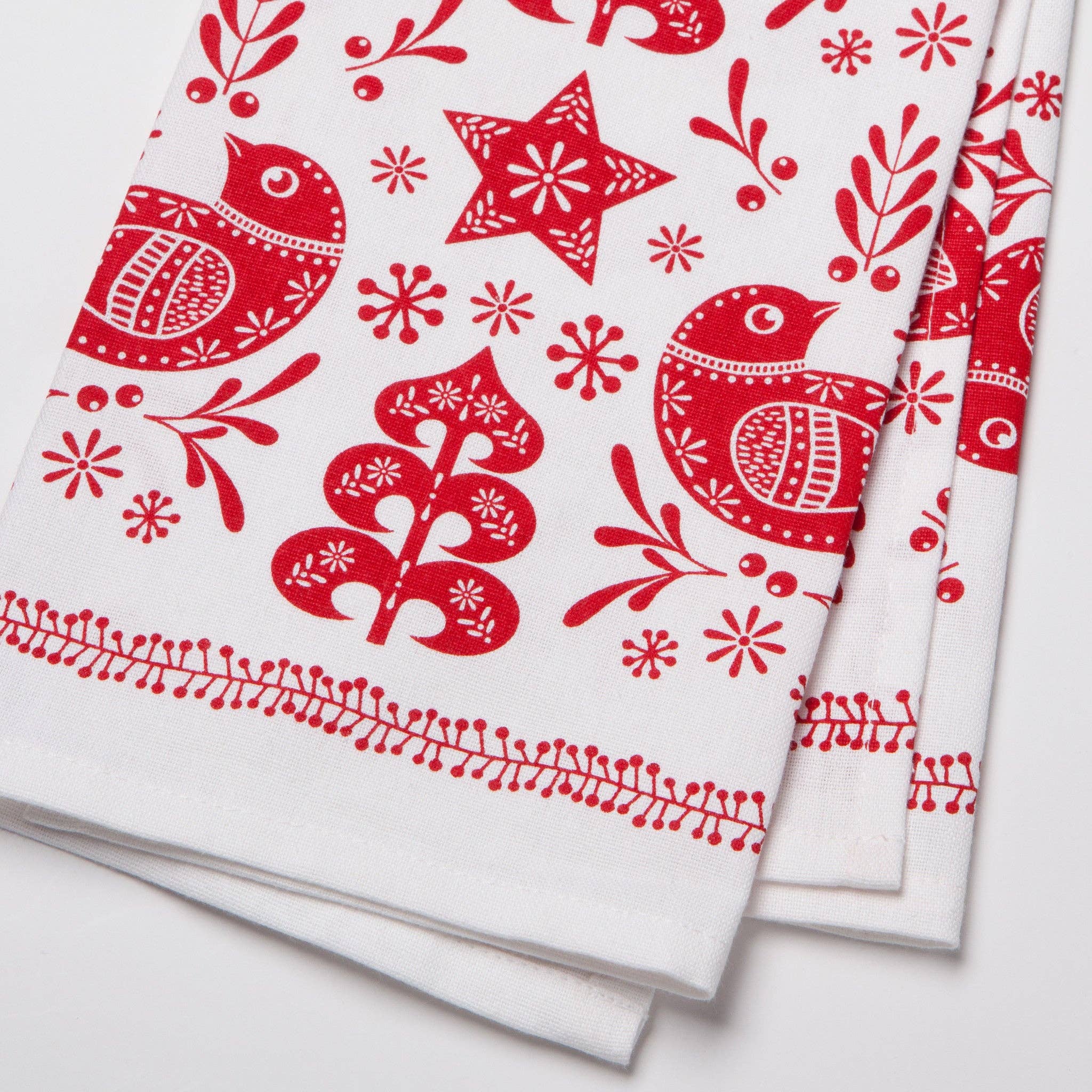 The "Tea Towel: Snowbird Dishtowel" is made from 100% cotton and features red Scandinavian-style illustrations of birds, stars, and trees, making it perfect for adding a touch of charm to your kitchen decor.