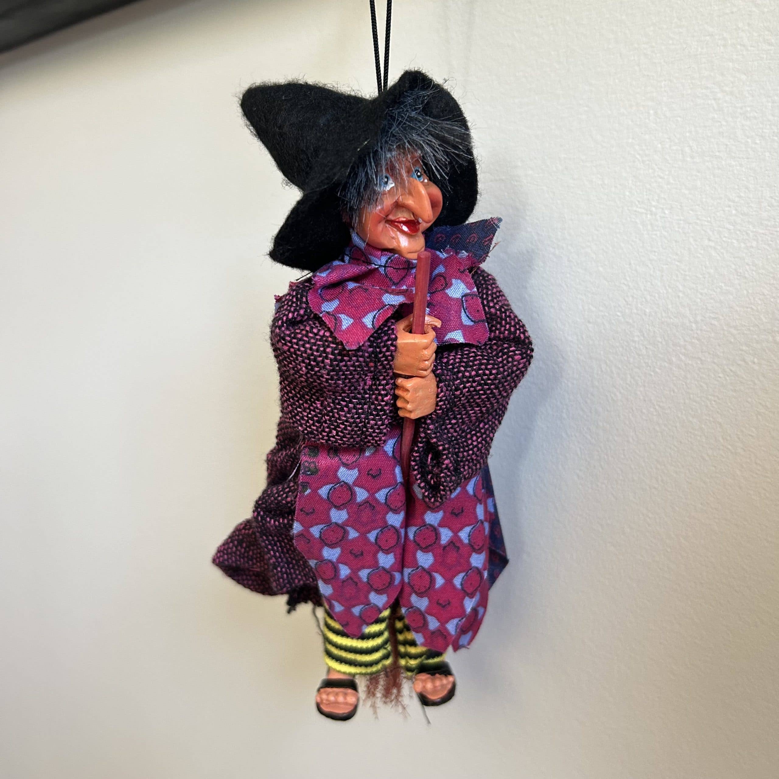 The Johanna Purple Kitchen Witch Doll Figurine, 9" tall, features a black hat, purple dress, and yellow-black striped socks while holding a broomstick—an adorable good luck charm to hang on your wall.