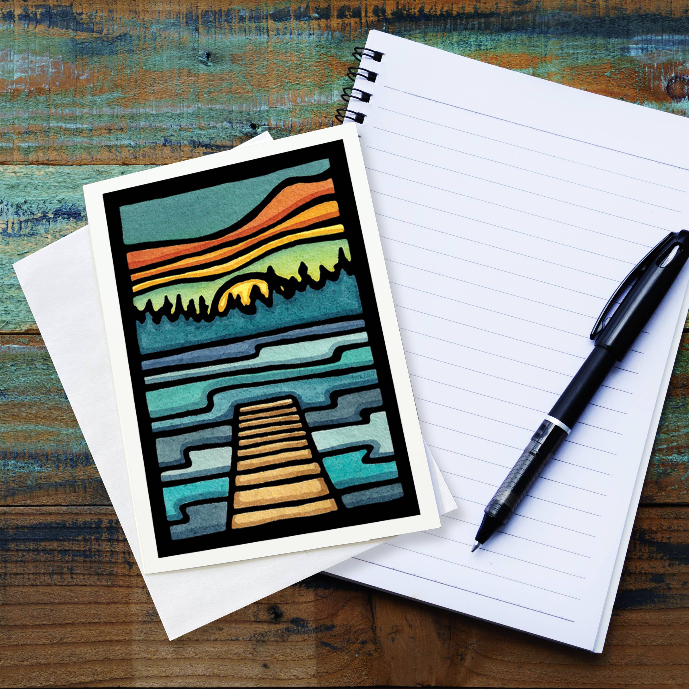 A vibrant greeting card titled "Lake Life Dock" by Sarah Angst is placed on a wooden table next to a blank notepad and a black pen, capturing the energetic style of Sarah Angst Art's linocut prints.