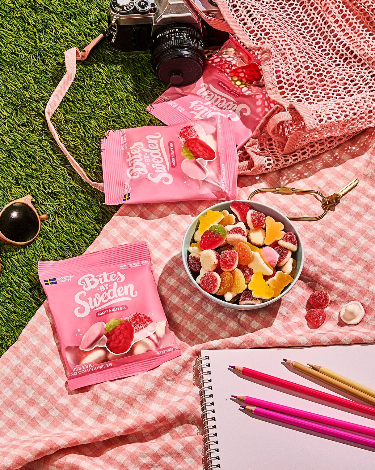 A bowl of assorted gummy candy with pink candy bags, colored pencils, a notebook, camera, and sunglasses on a pink gingham cloth atop artificial grass.