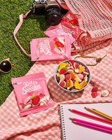 A bowl of assorted gummy candy with pink candy bags, colored pencils, a notebook, camera, and sunglasses on a pink gingham cloth atop artificial grass.