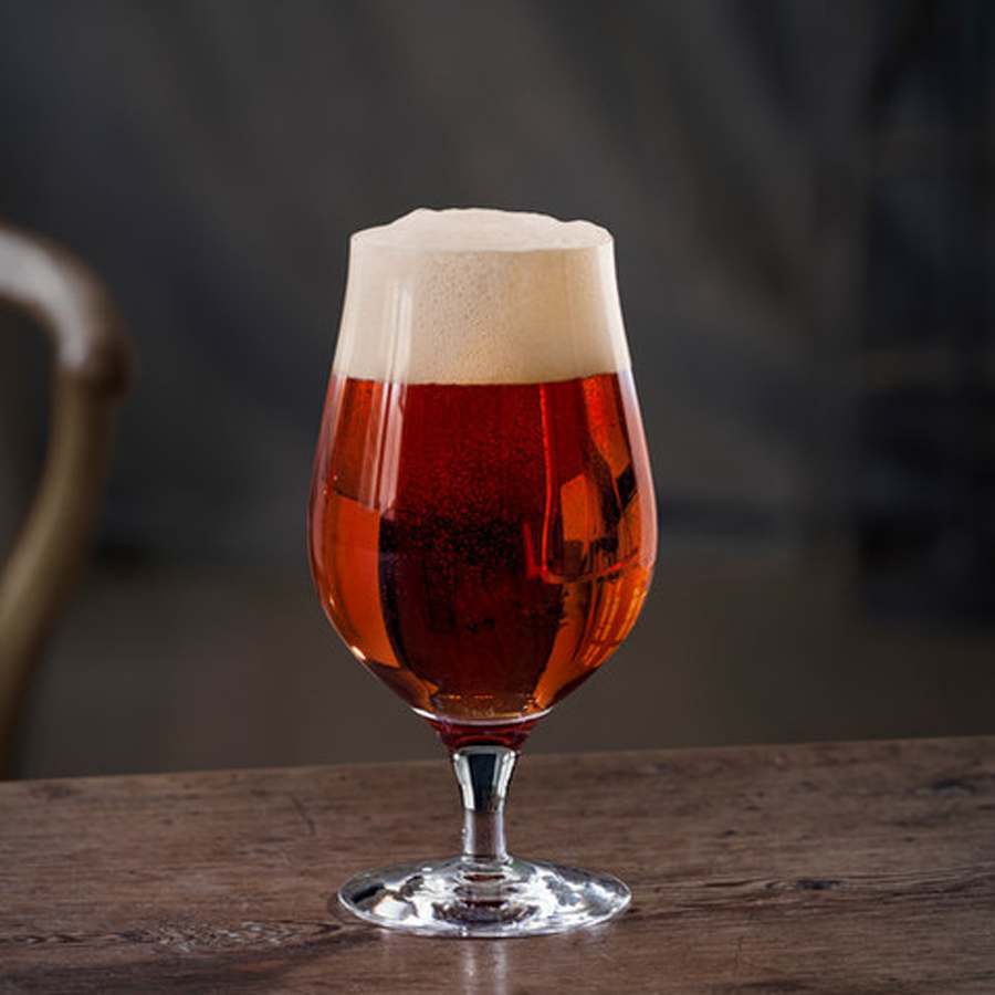 An Orrefors Beer Taster Glass 16oz from the 4-pack, filled with amber beer and a frothy head, sits elegantly on a wooden table.