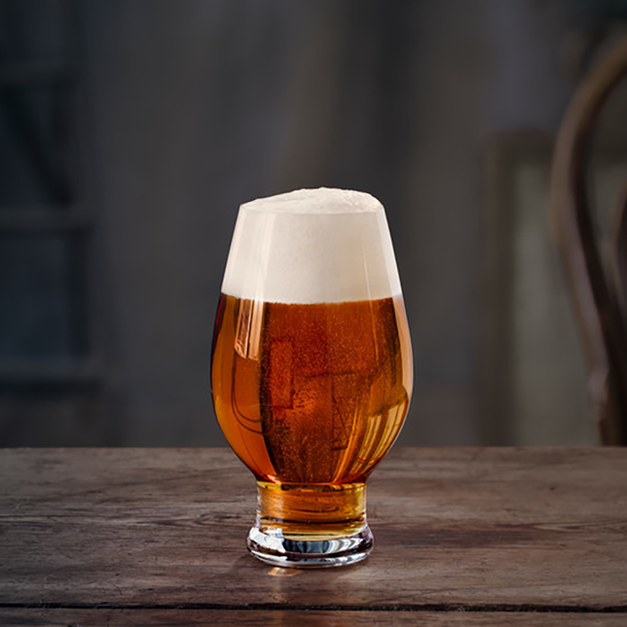 A glass of amber beer with a frothy head sits elegantly on a wooden table, perfectly cradled in an Orrefors: Beer IPA 15.5oz glass from the 4-Pack set.