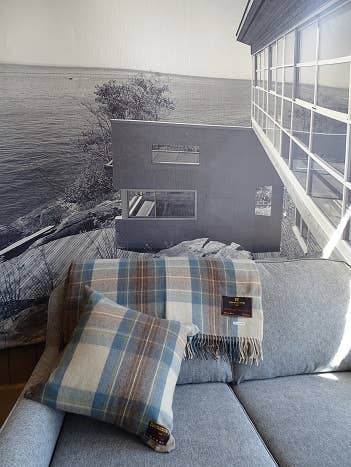 A gray sofa with the Stewart Muted Blue Deluxe Highland Blanket and a blue and beige pillow, set against a wall mural of a modern house by the water.