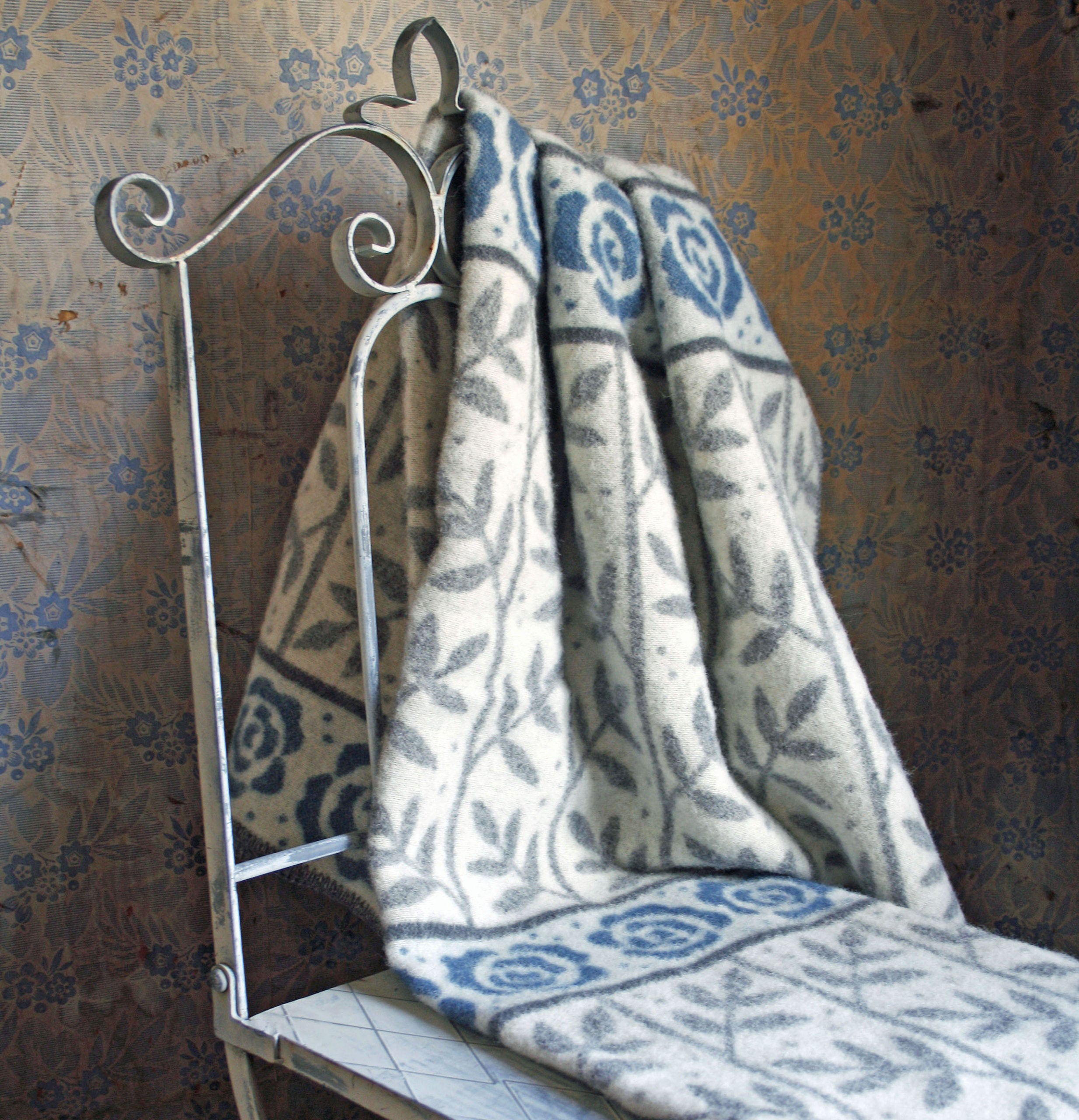 The Rose Garden Blanket by Kerstin Landstrom, made of 100% Norwegian Lambs Wool and OEKO-TEX-certified, elegantly drapes over a decorative metal chair, perfectly complementing the floral-patterned wallpaper.