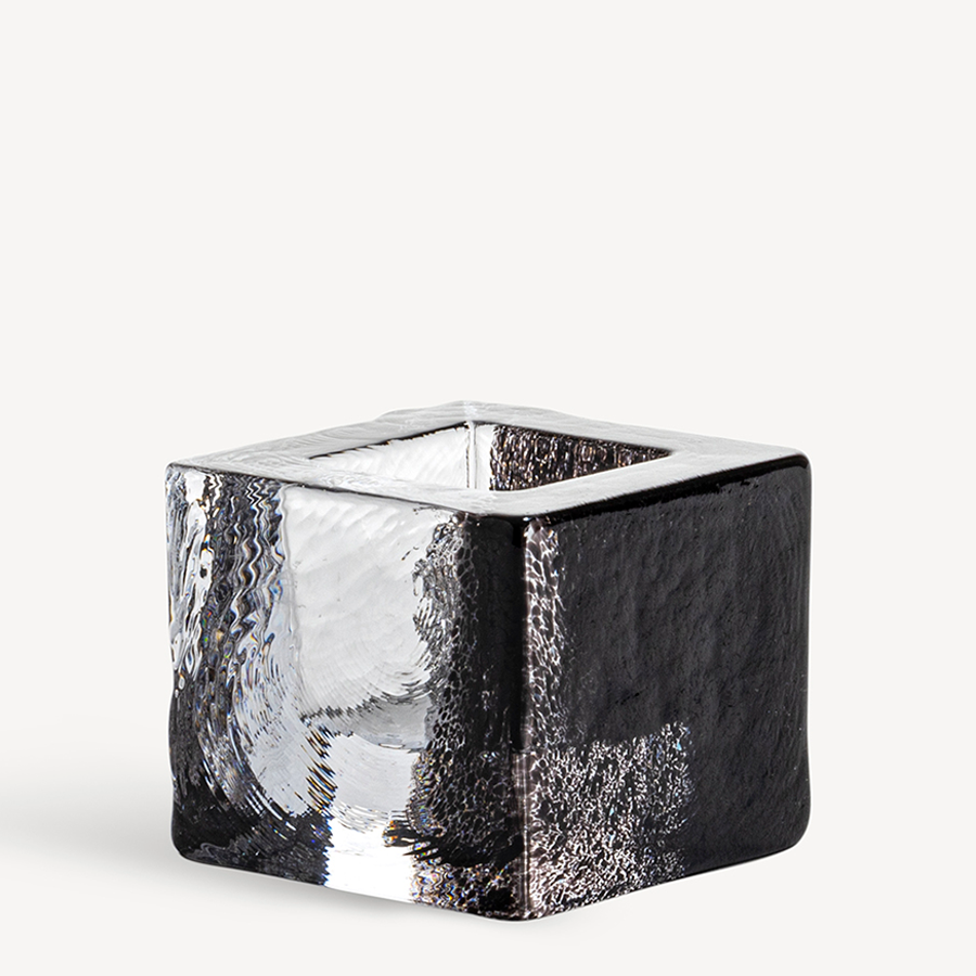 The KostaBoda Brick Votive Candle Holder features a textured black and clear design against a simple white backdrop. With echoes of Anna Ehrners style, it embodies the elegance and detailed craftsmanship that defines Kosta Boda, making it perfect for any setting.