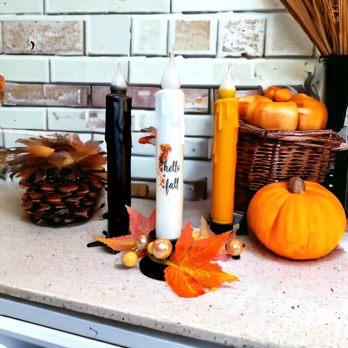 Three decorative candles with autumn leaves, pumpkins, and a pinecone on the counter. A white candle says Hello Fall. Enhance your farmhouse décor using the Hand-Dipped Beeswax 7 Tapered LED Light Candle featuring a battery-operated timer for ambiance.