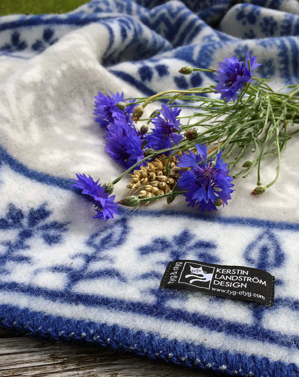 The blanket, named Blaklint Cornflower, is made from 100% Norwegian lambs wool. Designed by Kerstin Landstrom, it features a white and blue pattern with elegant blue flowers reminiscent of Blaklint cornflowers, adorned with small pine cones.