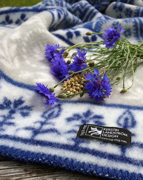 The blanket, named Blaklint Cornflower, is made from 100% Norwegian lambs wool. Designed by Kerstin Landstrom, it features a white and blue pattern with elegant blue flowers reminiscent of Blaklint cornflowers, adorned with small pine cones.