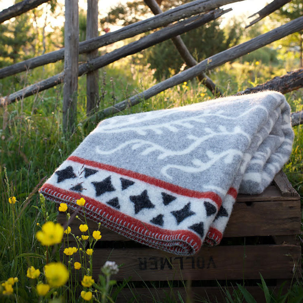 The Albertus Blanket made of 100% Norwegian lambs wool by Kerstin Landstrom, featuring stars and stripes, is draped over a wooden crate amidst yellow flowers with a wooden fence in the background.