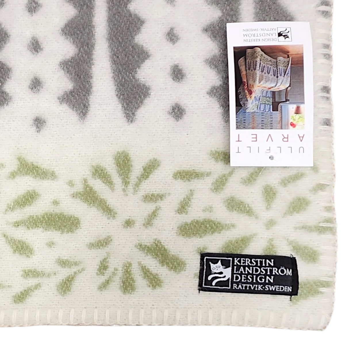 Patterned wool throw with green and gray designs, rectangular label, and product tag attached; Swedish design visible on soft woven surface.