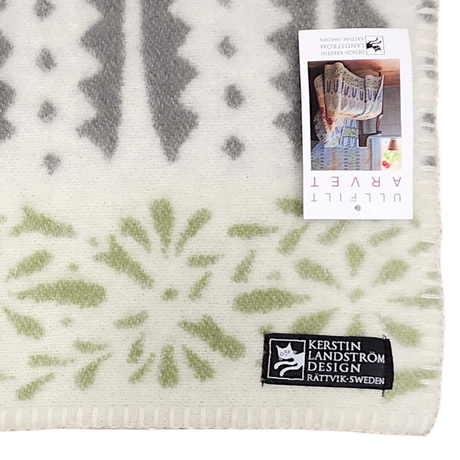 Patterned wool throw with green and gray designs, rectangular label, and product tag attached; Swedish design visible on soft woven surface.