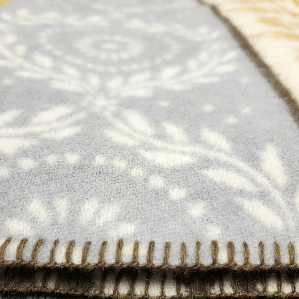 Closeup of a light gray lambswool blanket with a white floral pattern and dark stitched edge, partially draped over a yellow blanket with a geometric pattern underneath.