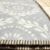 Closeup of a light gray lambswool blanket with a white floral pattern and dark stitched edge, partially draped over a yellow blanket with a geometric pattern underneath.