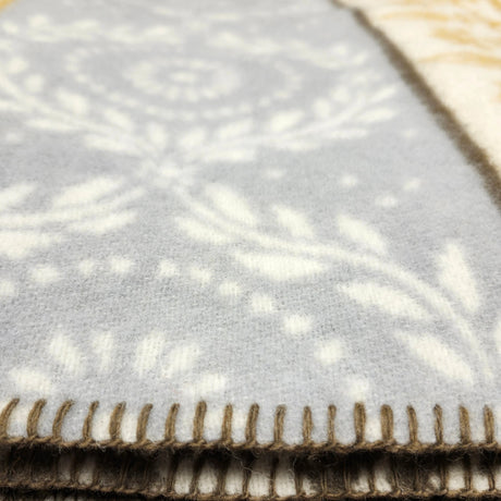 Closeup of a light gray lambswool blanket with a white floral pattern and dark stitched edge, partially draped over a yellow blanket with a geometric pattern underneath.