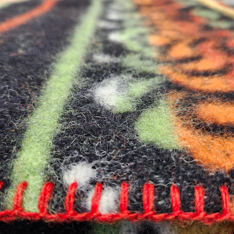Close-up of a lambswool blanket with a black, green, and orange pattern and visible red stitching along the edge.