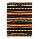 Striped Bunad blanket with black, red, yellow, and orange bands, detailed woven patterns, and a small label sewn onto the bottom right corner.