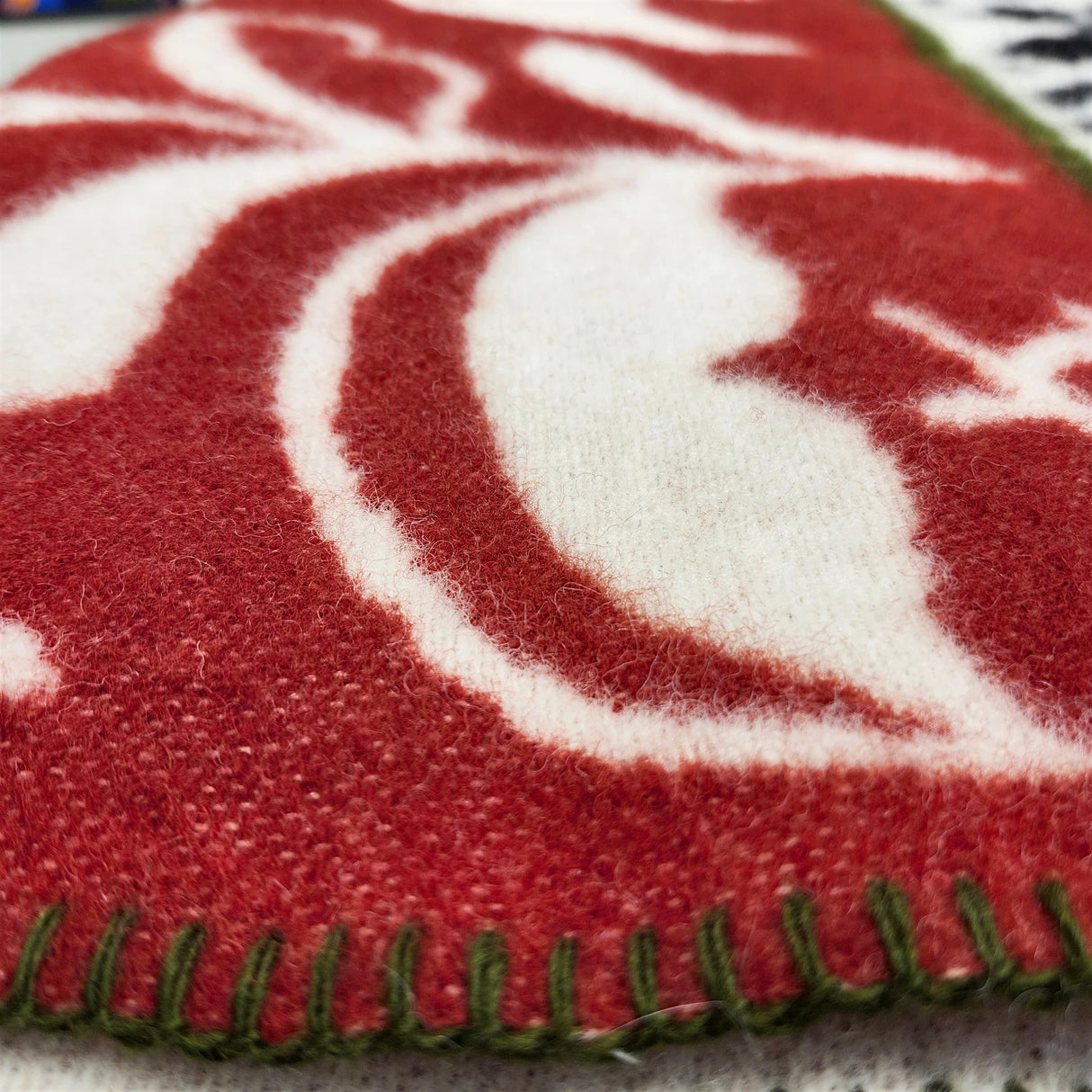 Close-up of a red and white patterned lambswool blanket with green edge stitching, showing detailed texture and fibers.