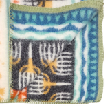 Close-up of a lambswool blanket featuring Swedish geometric patterns in blue, green, orange, and black, with green-stitched edges and one corner folded over.