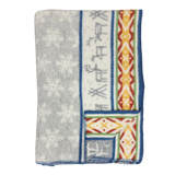 Folded Sámi-inspired wool blanket featuring a gray snowflake pattern, blue border, and one side with geometric and stripe designs in orange, yellow, and blue.