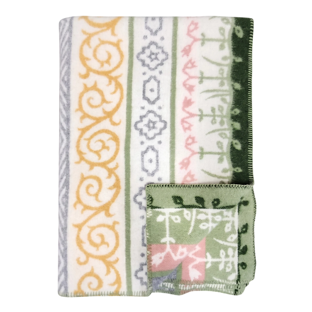 A folded blanket with green, yellow, gray, and pink geometric and floral patterns, plus a small OEKO-TEX certified square displaying matching designs on the edge.