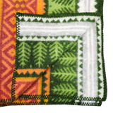 Close-up of a folded blanket featuring green, orange, and white geometric and leaf patterns, visible stitched edge, and soft lambswool texture.
