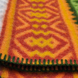 Close-up of a lambswool blanket featuring orange, yellow, and green geometric patterns with green-stitched edges in a Scandinavian design.