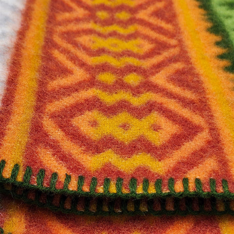 Close-up of a lambswool blanket featuring orange, yellow, and green geometric patterns with green-stitched edges in a Scandinavian design.