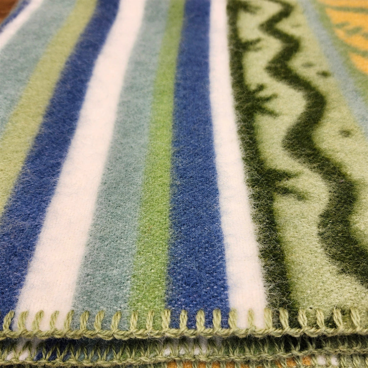 Close-up of a lambswool blanket with blue, green, and white stripes, Swedish design, decorative stitched edge, and wavy green patterns along one side.