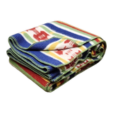 Folded wool blanket with colorful stripes and geometric patterns, green stitching along the edges. Scandinavian design elements visible.