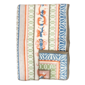 Folded wool blanket with blue, orange, green, and beige animal, floral, and geometric patterns inspired by Swedish folk art, brown trim visible along the edge.