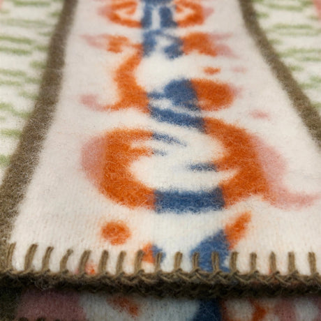 Close-up of a lambswool blanket with orange, blue, and white patterns and stitched edges, displayed on a surface featuring a green and white design.