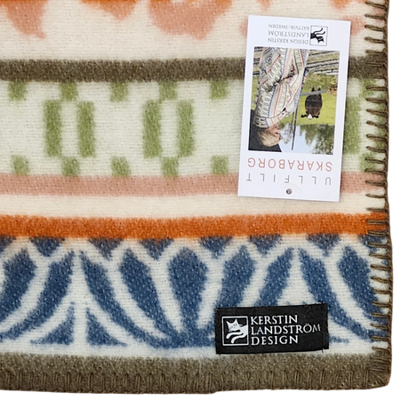 Patterned lambswool blanket with blue, orange, green, and white designs, Kerstin Landström Design label, and an upside-down product tag visible.
